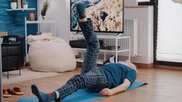 Uomo anziano sdraiato su un tappetino da yoga e allena i muscoli, solleva le gambe per fare esercizio fisico e allenamento. Persona anziana che fa attività di fitness con attrezzature sportive a casa. Stretching per adulti