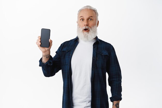 Uomo anziano sorpreso e impressionato che mostra lo schermo del telefono cellulare che tiene lo smartphone in mano ansimando e guardando stupito dalla fotocamera in piedi su sfondo bianco