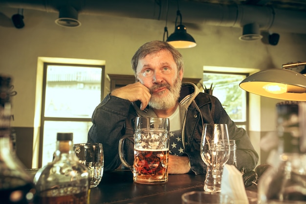 Uomo anziano triste che beve alcolici in un pub e guarda un programma sportivo in TV