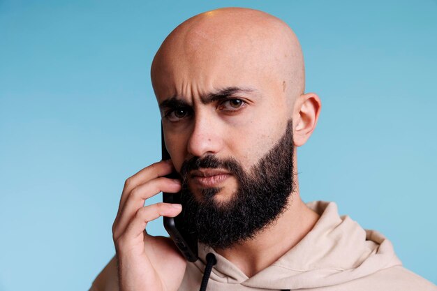Uomo arabo confuso che risponde alla chiamata dello smartphone e ritratto di sopracciglia accigliate. Giovane persona barbuta calva che parla al cellulare e guarda l'obbiettivo con espressione incerta
