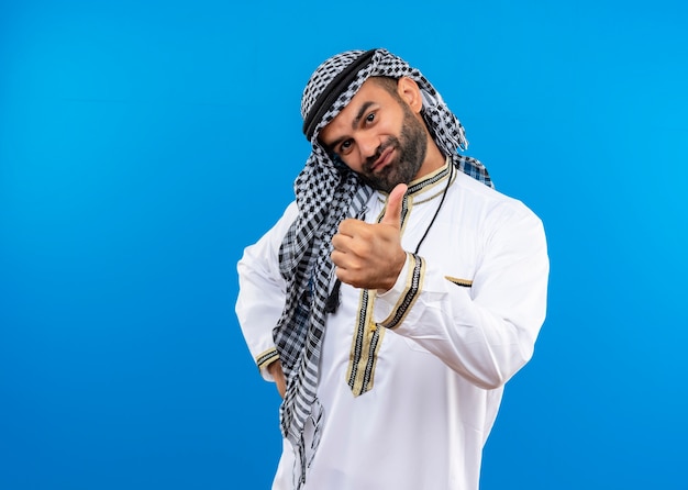 Uomo arabo in abbigliamento tradizionale che mostra i pollici in su con il sorriso sul viso in piedi sopra la parete blu