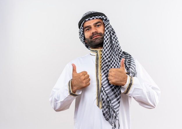 Uomo arabo in usura tradizionale sorridente che mostra i pollici in su in piedi sul muro bianco