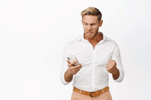 Uomo arrabbiato che guarda il cellulare con espressione di faccia furiosa incazzata frustrato per qualcosa sul telefono in piedi su sfondo bianco