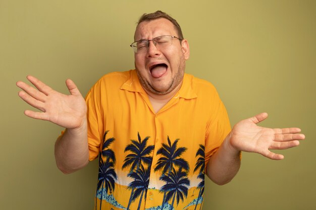 Uomo arrabbiato con gli occhiali che indossa la camicia arancione che grida e urla con le braccia alzate in piedi sopra il muro verde