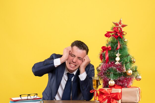 Uomo arrabbiato di vista frontale che tiene la sua testa che si siede al tavolo vicino all'albero di Natale e regali su priorità bassa gialla