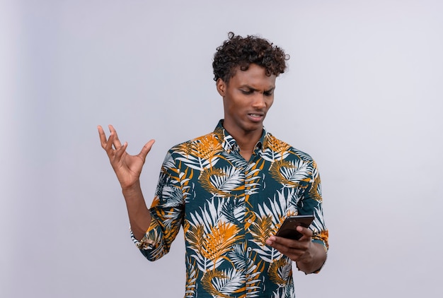 Uomo arrabbiato e nervoso dalla carnagione scura in camicia stampata foglie guardando il cellulare e alzando la mano