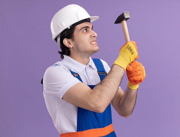 Uomo arrabbiato giovane costruttore in uniforme da costruzione e casco di sicurezza in guanti di gomma che tiene martello guardandolo con espressione infastidita in piedi sopra la parete viola