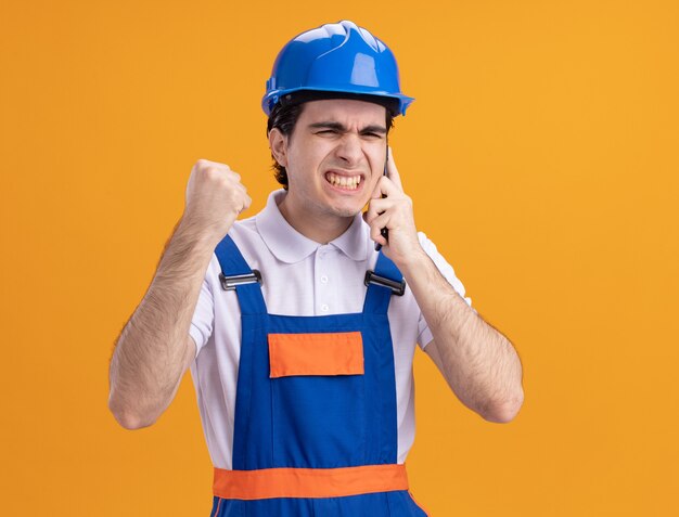 Uomo arrabbiato giovane costruttore in uniforme da costruzione e casco di sicurezza parlando al telefono cellulare in piedi sopra la parete arancione