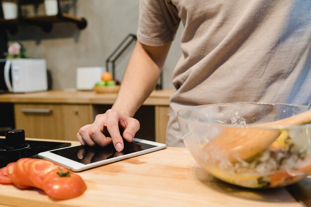 Uomo asiatico che guarda ricetta sulla compressa digitale e che cucina alimento sano nella cucina domestica