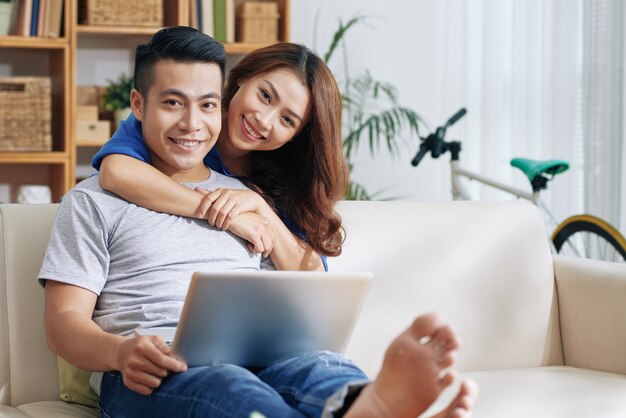 Uomo asiatico che si rilassa sullo strato con il computer portatile a casa e la donna felice che lo abbraccia