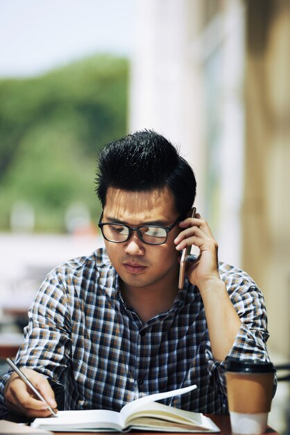 Uomo asiatico che si siede in caffè all'aperto, parlando sul telefono cellulare e scrivere nel quaderno