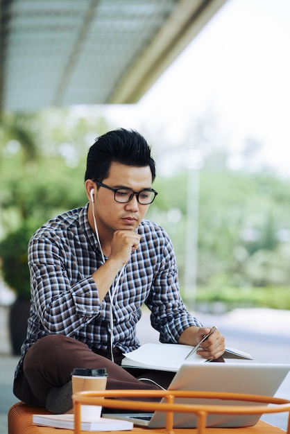 Uomo asiatico che si siede sul banco all'aperto con il computer portatile e che ascolta il webinar online