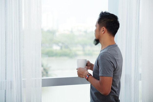 Uomo asiatico che sta davanti alla finestra alta con la tazza e che guarda fuori