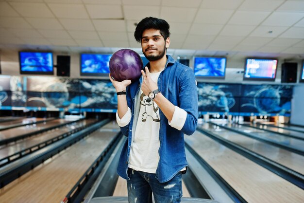 Uomo asiatico con barba alla moda in camicia di jeans in piedi alla pista da bowling con palla a portata di mano