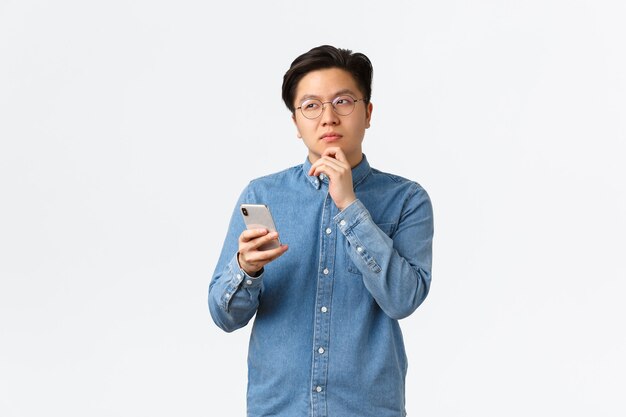 Uomo asiatico creativo premuroso con gli occhiali che pensa mentre pubblica post sui social media, distoglie lo sguardo, medita o prende una decisione, tiene in mano uno smartphone, sceglie qualcosa su internet.