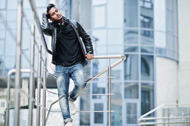 Uomo asiatico elegante e casual in giacca di pelle nera posato vicino alla ringhiera contro un edificio moderno