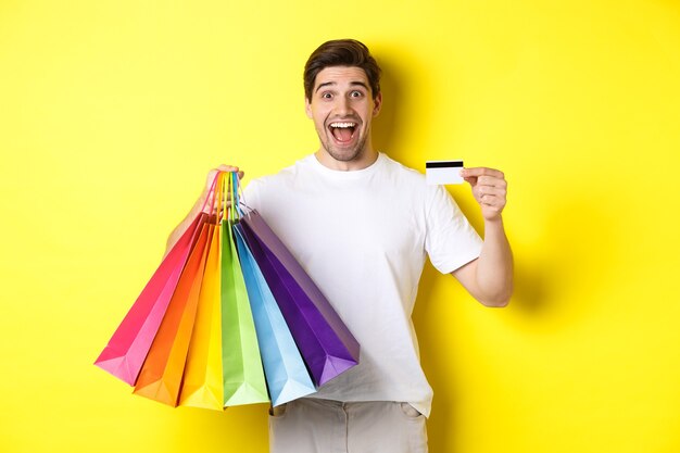 Uomo attraente felice che tiene le borse della spesa, mostrando la carta di credito, in piedi su sfondo giallo
