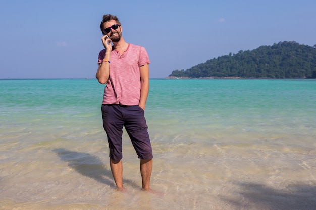 Uomo attraente felice in occhiali da sole alla moda con la barba utilizzando il telefono cellulare in spiaggia