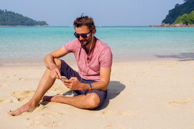 Uomo attraente felice in occhiali da sole alla moda con la barba utilizzando il telefono cellulare in spiaggia