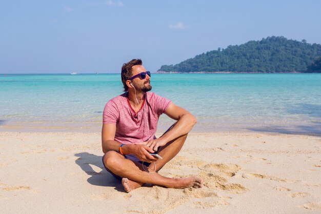 Uomo attraente felice in occhiali da sole alla moda con la barba utilizzando il telefono cellulare in spiaggia