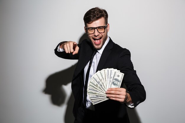 Uomo attraente felice in vetri e vestito nero che tiene mazzo di soldi mentre indicando con il dito su voi