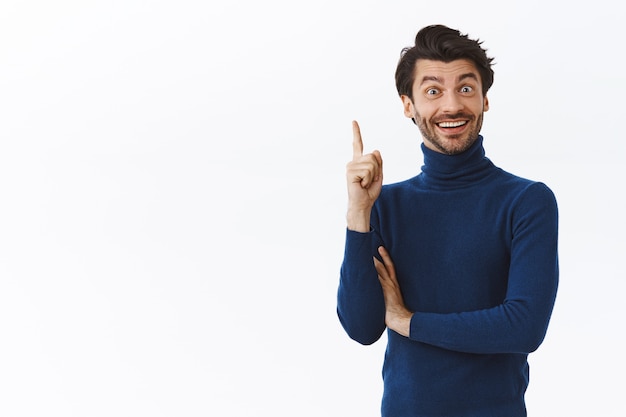 Uomo attraente in elegante maglione a collo alto, alza il dito indice nel gesto di eureka sorridendo con gioia, trova la risposta, dà un suggerimento interessante, felice finalmente risolve il problema, muro bianco