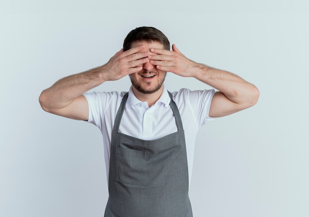 uomo barbiere in grembiule che copre gli occhi con le braccia sorridenti in piedi sul muro bianco