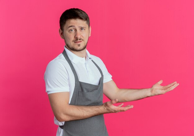 uomo barbiere in grembiule che presenta qualcosa con le braccia sorridente in piedi fiducioso sopra il muro rosa