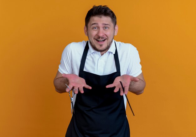 uomo barbiere in grembiule con forbici e pettine confuso sorridente in piedi sopra il muro arancione