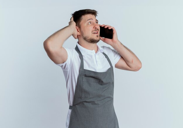 uomo barbiere in grembiule guardando confuso mentre parla al telefono cellulare in piedi sopra il muro bianco