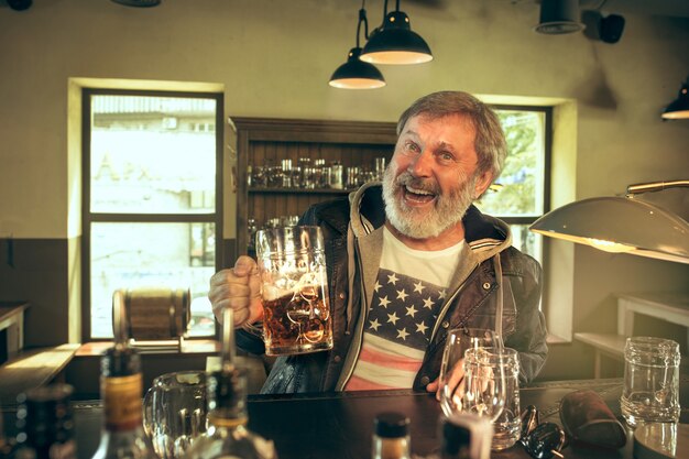 Uomo barbuto anziano che beve alcolici in un pub e guarda un programma sportivo in TV. Gustando il mio brulicare e la mia birra preferiti. Uomo con boccale di birra seduto a tavola. Appassionato di calcio o di sport. Concetto di emozioni umane