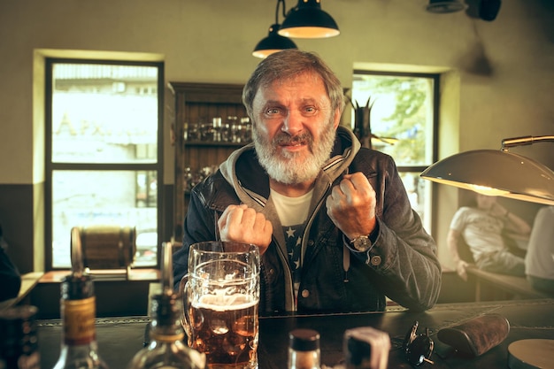 Uomo barbuto anziano che beve alcolici in un pub e guarda un programma sportivo in TV. Gustando il mio brulicare e la mia birra preferiti. Uomo con boccale di birra seduto a tavola.
