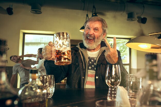 Uomo barbuto anziano che beve alcolici in un pub e guarda un programma sportivo in TV.