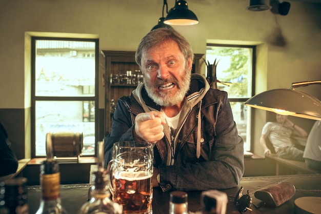 Uomo barbuto arrabbiato che beve alcolici nel pub e guarda un programma sportivo in Tv. Gustando il mio brulicare e la mia birra preferiti. Uomo con boccale di birra seduto a tavola. Appassionato di calcio o di sport. Concetto di emozioni umane