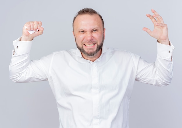 Uomo barbuto arrabbiato che indossa una camicia bianca che si scatena alzando le mani in piedi sul muro bianco