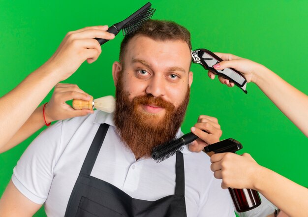 Uomo barbuto barbiere in grembiule sorridente avendo il suo taglio di capelli e la sua barba modellata da altre mani sul muro verde