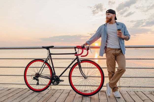 Uomo barbuto bello che viaggia con la bicicletta nell'alba di mattina in riva al mare a bere caffè, viaggiatore sano stile di vita attivo