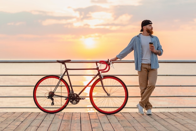 Uomo barbuto bello che viaggia con la bicicletta nell'alba di mattina in riva al mare a bere caffè, viaggiatore sano stile di vita attivo