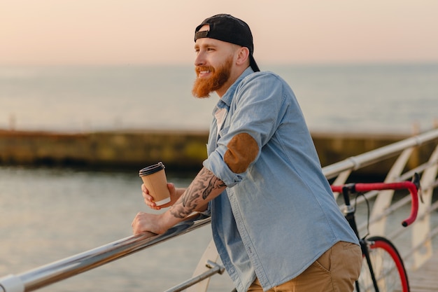 Uomo barbuto bello che viaggia con la bicicletta nell'alba di mattina in riva al mare a bere caffè, viaggiatore sano stile di vita attivo