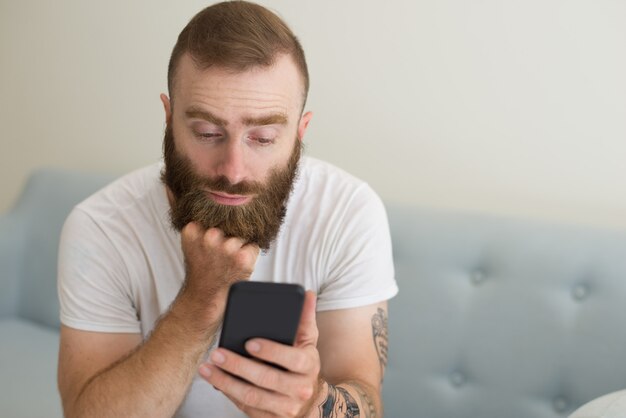 Uomo barbuto bello noioso che utilizza smartphone nel salone