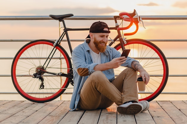 Uomo barbuto bello stile hipster che indossa camicia di jeans e cappuccio che tiene smartphone con la bicicletta all'alba di mattina in riva al mare, bere caffè, viaggiatore sano stile di vita attivo