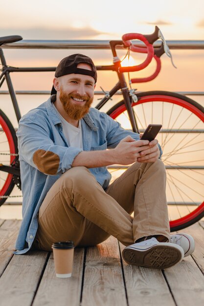 Uomo barbuto bello stile hipster che indossa camicia di jeans e cappuccio che tiene smartphone con la bicicletta all'alba di mattina in riva al mare, bere caffè, viaggiatore sano stile di vita attivo