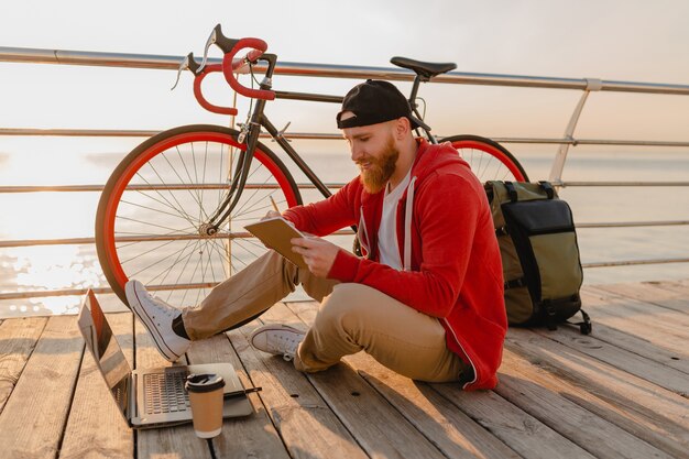 Uomo barbuto bello stile hipster che lavora libero professionista online su laptop con zaino e bicicletta in alba di mattina in riva al mare viaggiatore zaino in spalla sano stile di vita attivo