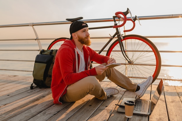 Uomo barbuto bello stile hipster che lavora libero professionista online su laptop con zaino e bicicletta in alba di mattina in riva al mare viaggiatore zaino in spalla sano stile di vita attivo