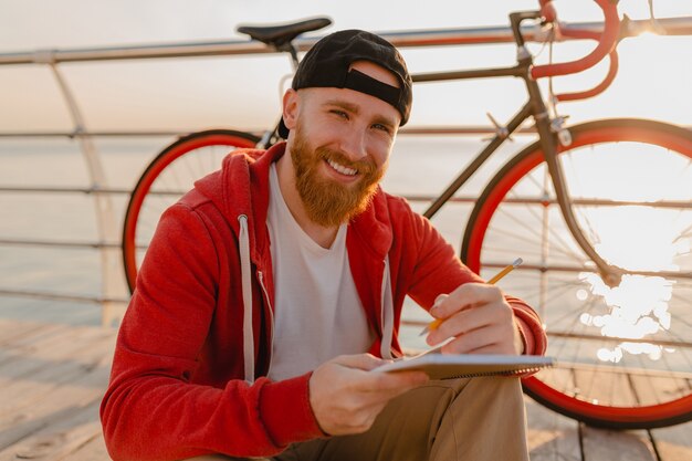 Uomo barbuto bello stile hipster che lavora libero professionista online su laptop con zaino e bicicletta in alba di mattina in riva al mare viaggiatore zaino in spalla sano stile di vita attivo