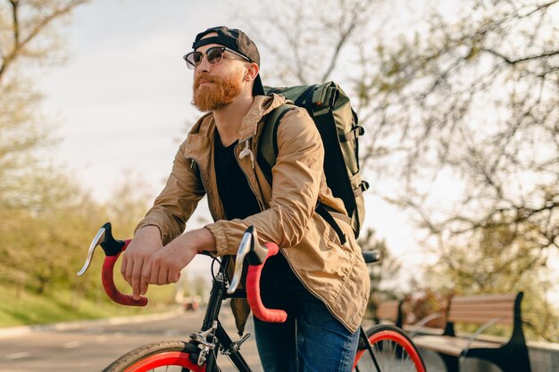 Uomo barbuto bello stile hipster in giacca e occhiali da sole in sella da solo con lo zaino in bicicletta viaggiatore zaino in spalla sano stile di vita attivo