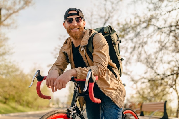 Uomo barbuto bello stile hipster in giacca e occhiali da sole in sella da solo con lo zaino in bicicletta viaggiatore zaino in spalla sano stile di vita attivo