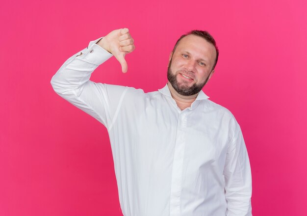 Uomo barbuto che indossa una camicia bianca cercando dispiaciuto che mostra i pollici verso il basso in piedi sul muro rosa