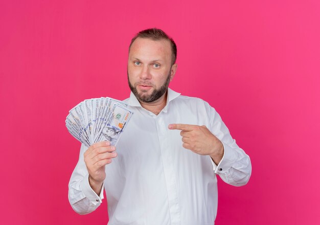 Uomo Barbuto che indossa una camicia bianca che mostra denaro contante puntato con il dito indice al denaro cercando fiducioso in piedi oltre il muro rosa