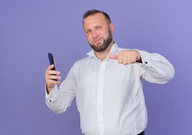 Uomo Barbuto che indossa una camicia bianca che mostra lo smartphone che punta il dito contro sorridente fiducioso in piedi oltre la parete blu